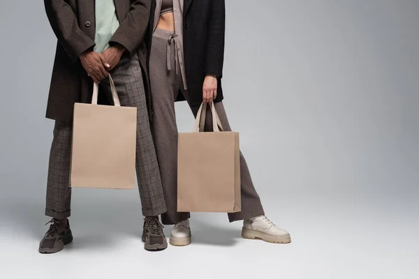 Vista recortada de elegante pareja interracial en trajes otoñales sosteniendo bolsas de compras en gris - foto de stock