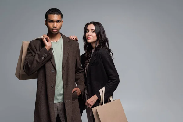 Stylish interracial couple in autumnal outfits holding shopping bags isolated on grey — Stock Photo