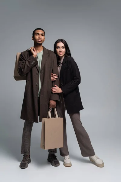 Full length of stylish interracial couple in trendy autumnal outfits holding shopping bags on grey — Stock Photo