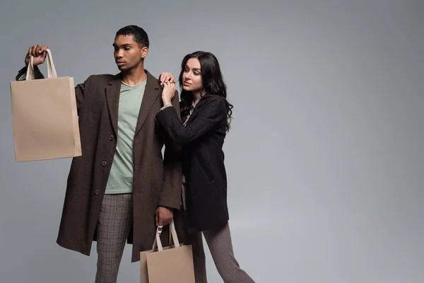 Mujer con estilo en blazer negro cerca de hombre joven afroamericano en abrigo sosteniendo bolsas de compras aisladas en gris - foto de stock