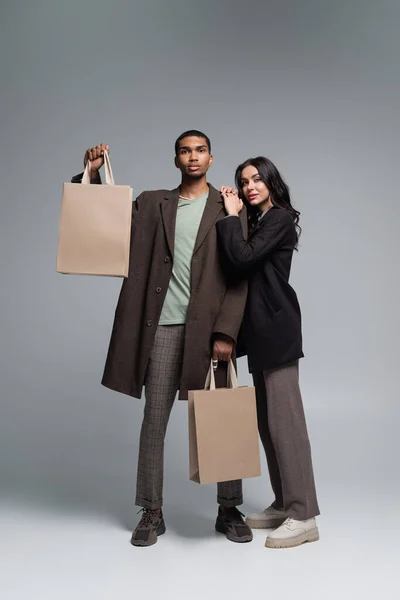 Full length of stylish interracial couple in autumnal outfits holding paper bags on grey — Stock Photo