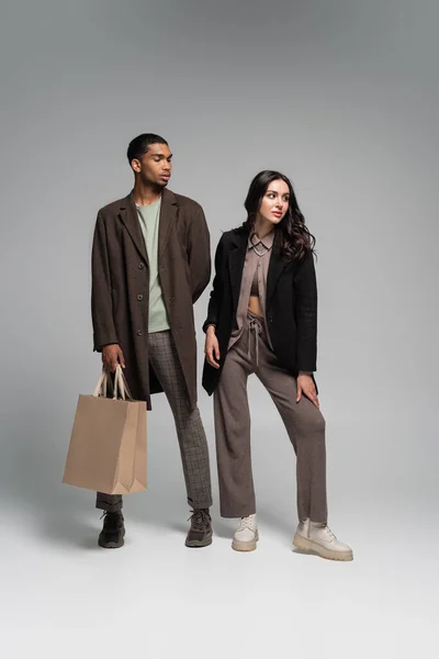 Longitud completa de elegante afroamericano en capa otoñal posando con bolsas de papel cerca de la mujer en gris - foto de stock