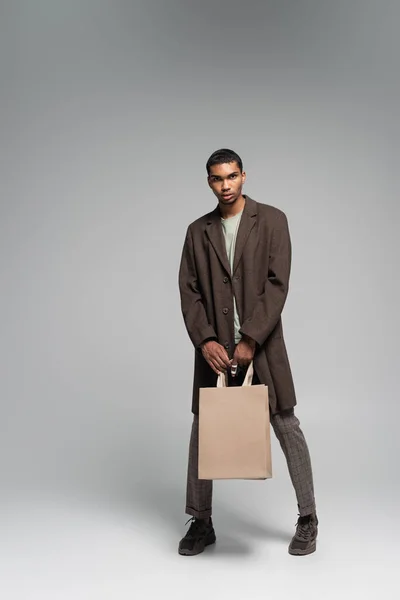 Pleine longueur de élégant homme afro-américain en manteau d'automne et baskets posant avec des sacs à provisions sur gris — Photo de stock