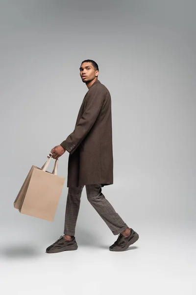 Piena lunghezza di elegante uomo afroamericano in cappotto autunnale e scarpe da ginnastica che camminano con shopping bag su grigio — Foto stock