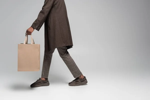 Cropped view of stylish african american man in autumnal coat and sneakers walking with shopping bags on grey — Stock Photo