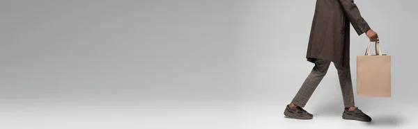Vista ritagliata di elegante uomo afroamericano in cappotto e scarpe da ginnastica che cammina con shopping bag su grigio, banner — Stock Photo