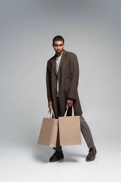 Comprimento total de homem americano africano elegante em casaco da moda e tênis posando com sacos de compras em cinza — Fotografia de Stock