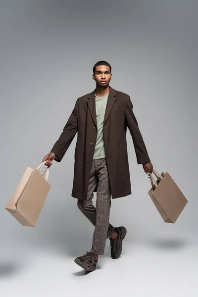 Comprimento total de homem americano africano elegante em casaco de lã e tênis posando com sacos de compras em cinza — Fotografia de Stock