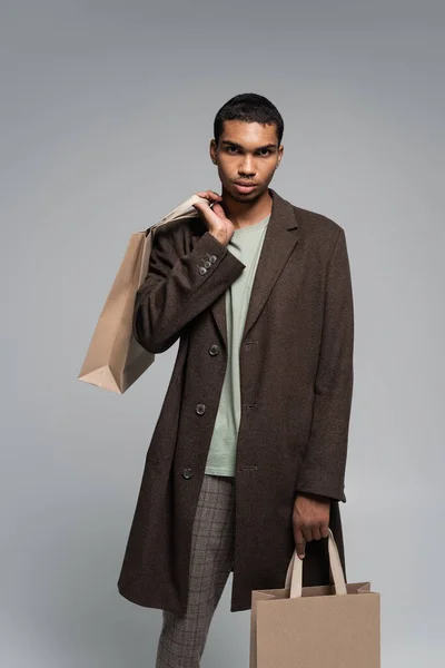 Young african american man in autumnal coat posing with shopping bags isolated on grey — Stock Photo