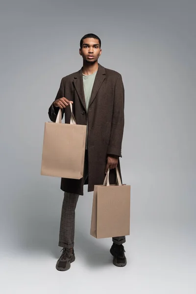 Full length of stylish african american man in autumnal coat and sneakers standing with shopping bags on grey — Stock Photo