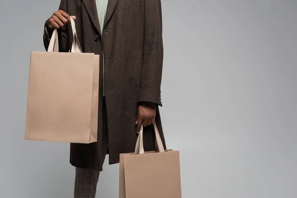 Vista recortada de hombre afroamericano con estilo en abrigo otoñal posando con bolsas de compras aisladas en gris - foto de stock