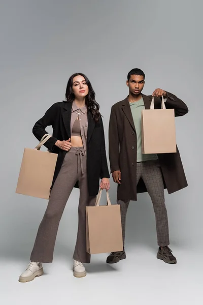 Longitud completa de elegantes modelos interracial en trajes otoñales sosteniendo bolsas de compras en gris - foto de stock