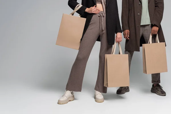 Vista parcial de elegante pareja interracial en trajes otoñales sosteniendo bolsas de compras en gris - foto de stock