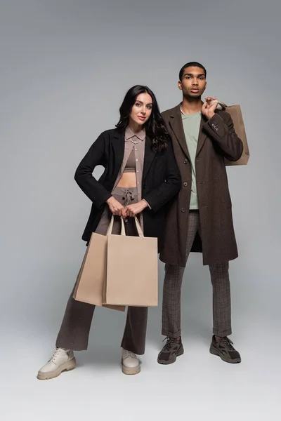 Toute la longueur du couple interracial à la mode dans des tenues élégantes tenant des sacs à provisions sur gris — Photo de stock