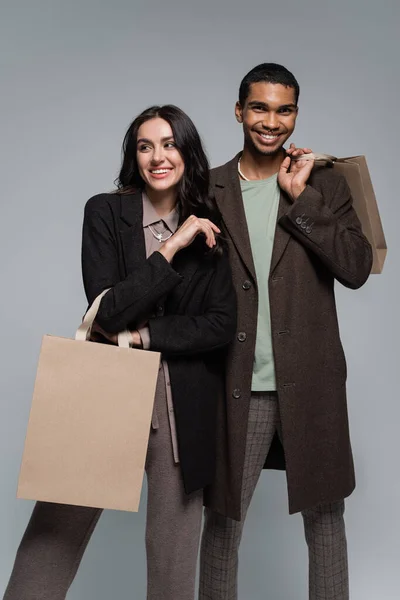 Feliz pareja interracial en elegantes trajes sosteniendo bolsas de compras aisladas en gris - foto de stock