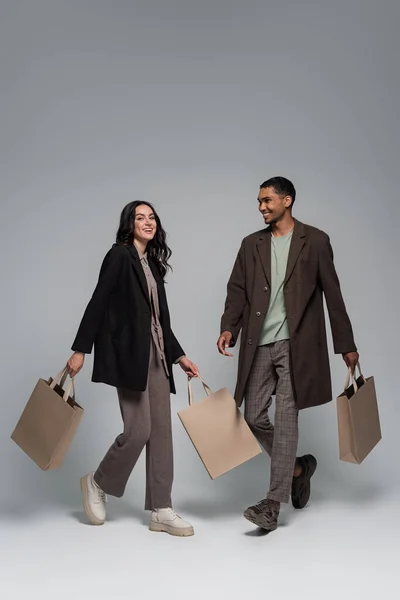 Longitud completa de feliz pareja interracial en trajes elegantes sosteniendo bolsas de compras y caminando en gris - foto de stock