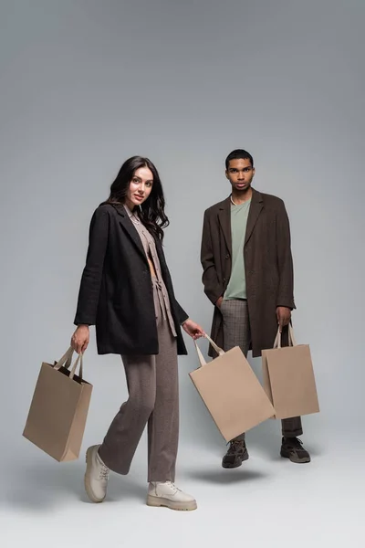 Full length of interracial couple in stylish outfits holding shopping bags and walking on grey — Stock Photo