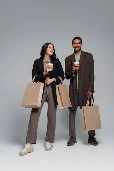 Longitud completa de feliz pareja interracial en trajes elegantes sosteniendo bolsas de compras y vasos de papel en gris - foto de stock