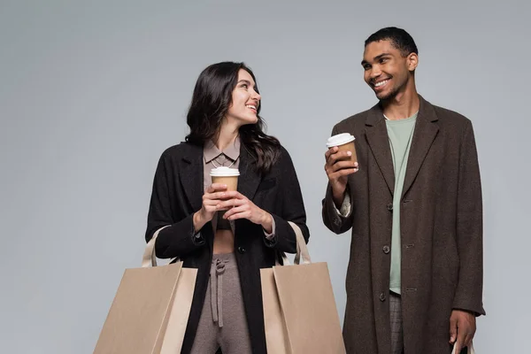 Happy interracial couple in stylish outfits holding shopping bags and paper cups isolated on grey — Stock Photo