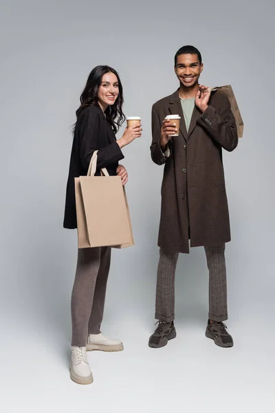 Full length of cheerful interracial couple in stylish outfits holding shopping bags and paper cups on grey — Stock Photo