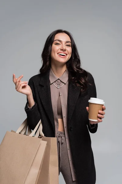 Happy young woman holding paper cup with coffee to go and shopping bags isolated on grey — Stock Photo