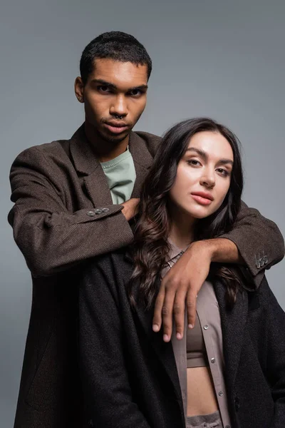 Africano americano hombre abrazando morena mujer en chaqueta aislado en gris - foto de stock