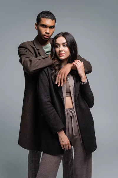 African american man hugging brunette woman in blazer and knitted trousers isolated on grey — Stock Photo