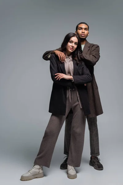 Full length of african american man hugging brunette woman in blazer isolated on grey — Stock Photo