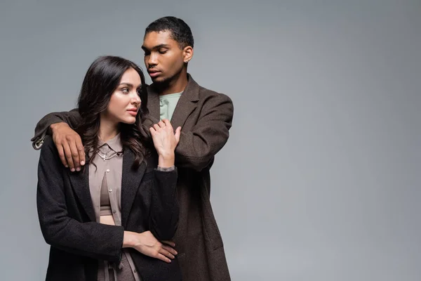 Young african american man hugging brunette woman in blazer isolated on grey — Stock Photo