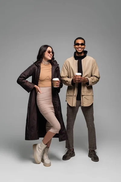 Pleine longueur de couple interracial gai dans des tenues élégantes et des lunettes de soleil tenant des tasses en papier sur gris — Photo de stock