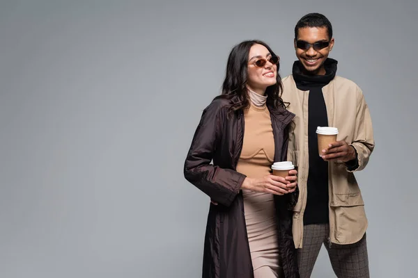 Alegre interracial casal no elegante roupas e óculos de sol segurando copos de papel isolado no cinza — Fotografia de Stock