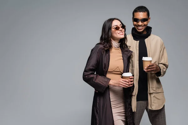 Alegre pareja interracial en elegantes trajes y gafas de sol sosteniendo vasos de papel aislados en gris - foto de stock