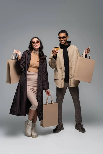 Comprimento total de modelos inter-raciais felizes em óculos de sol elegantes segurando sacos de compras e cartão de crédito em cinza — Fotografia de Stock