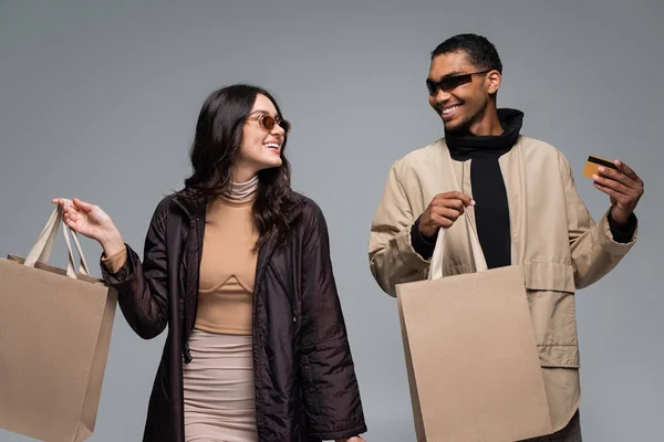 Heureux modèles interracial dans des lunettes de soleil élégantes tenant des sacs à provisions et carte de crédit isolé sur gris — Photo de stock