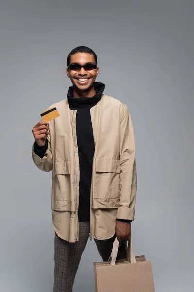 Felice uomo africano americano in eleganti occhiali da sole e vestito autunnale con carta di credito isolata su grigio — Foto stock
