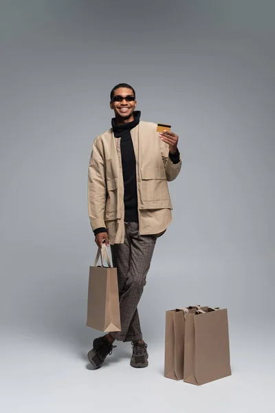 Piena lunghezza di felice uomo africano americano in occhiali da sole e vestito autunnale con carta di credito e shopping bag su grigio — Foto stock