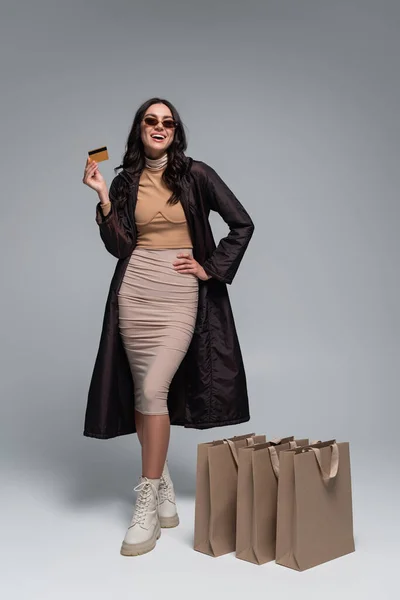 Piena lunghezza di giovane donna sorridente in abito elegante in possesso di carta di credito vicino shopping bags su grigio — Foto stock