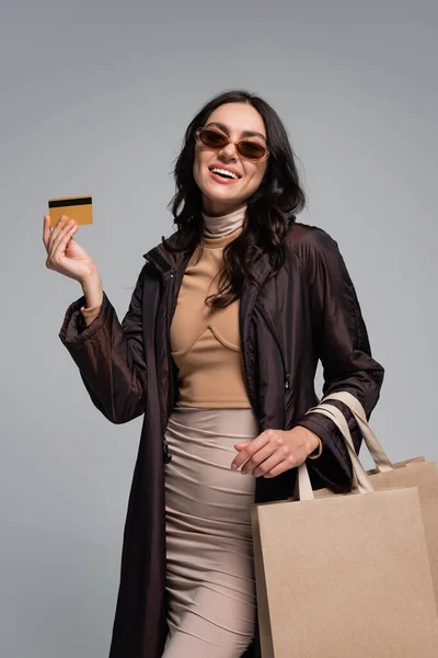 Усміхнена молода жінка в стильних сонцезахисних окулярах тримає сумки і кредитну картку ізольовано на сірому — стокове фото
