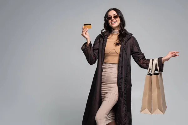 Smiling young woman in stylish sunglasses holding shopping bags and credit card isolated on grey — Stock Photo