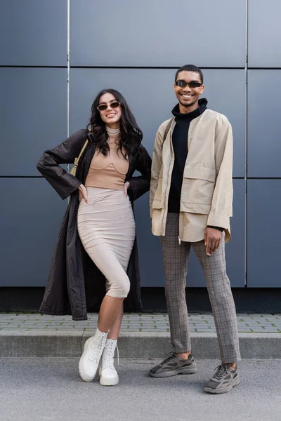 Полная длина стильной межрасовой пары в осенних нарядах и солнцезащитных очках, стоящих рядом со зданием — стоковое фото