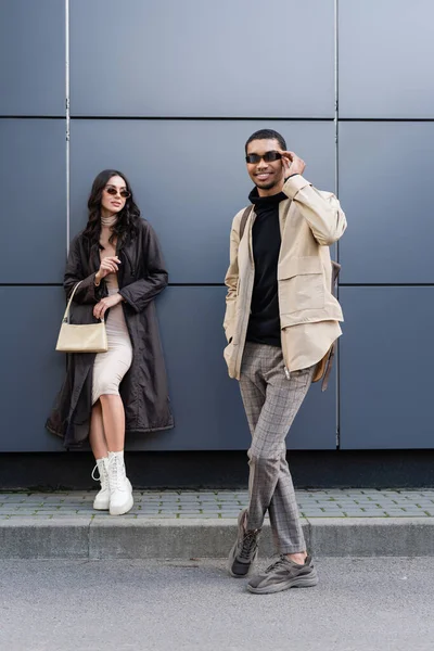 Piena lunghezza di felice uomo africano americano regolazione occhiali da sole vicino donna elegante in piedi con borsa — Foto stock