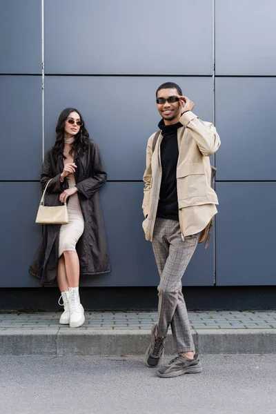 Comprimento total de homem americano africano feliz ajustando óculos de sol perto de mulher elegante de pé com bolsa — Fotografia de Stock