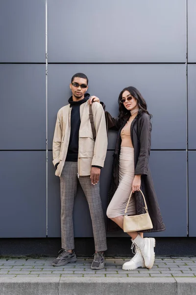 Full length of stylish interracial couple in autumnal outfits and trendy sunglasses standing near building — Stock Photo