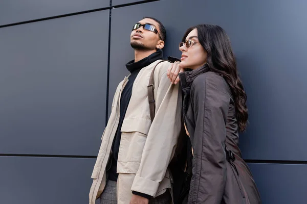 Pareja interracial con estilo en trajes otoñales y gafas de sol de moda de pie cerca del edificio - foto de stock