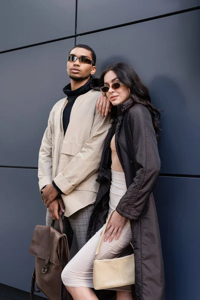 Homme afro-américain en lunettes de soleil tenant sac à dos en cuir près de femme élégante et bâtiment — Photo de stock