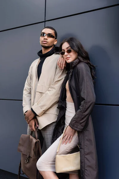 Africano americano hombre en gafas de sol celebración de cuero mochila cerca de la mujer con estilo y la construcción - foto de stock