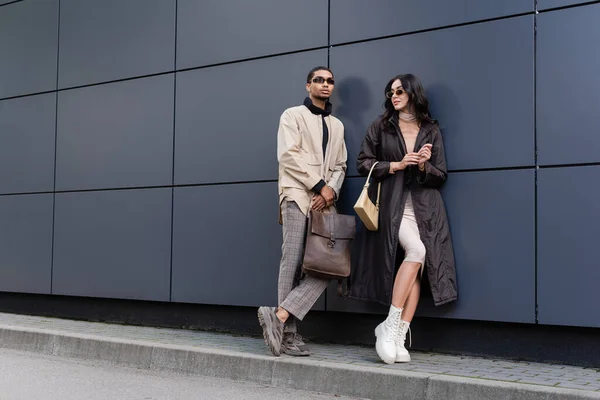 Longitud completa de hombre afroamericano en gafas de sol que sostienen la mochila de cuero cerca de la mujer con estilo con bolso - foto de stock