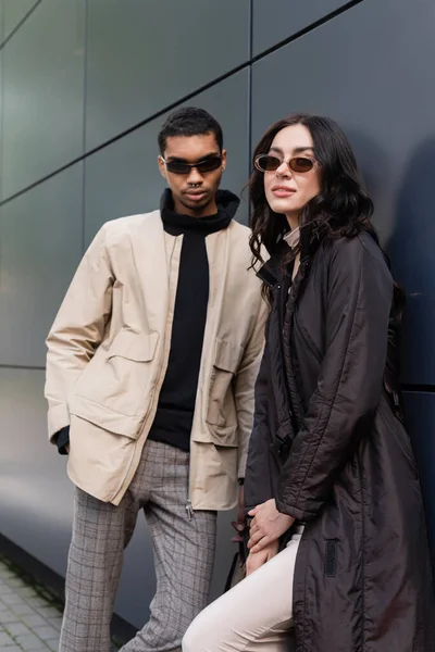 Elegante uomo africano americano in occhiali da sole in piedi con mano in tasca vicino alla giovane donna in cappotto — Foto stock