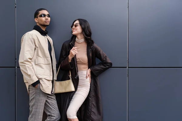 Élégant homme afro-américain en lunettes de soleil debout avec la main dans la poche près de jeune femme heureuse dans le manteau — Photo de stock