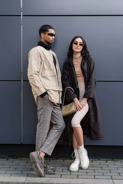 Hombre afroamericano con estilo en gafas de sol de pie con la mano en el bolsillo cerca de la mujer joven con bolso - foto de stock